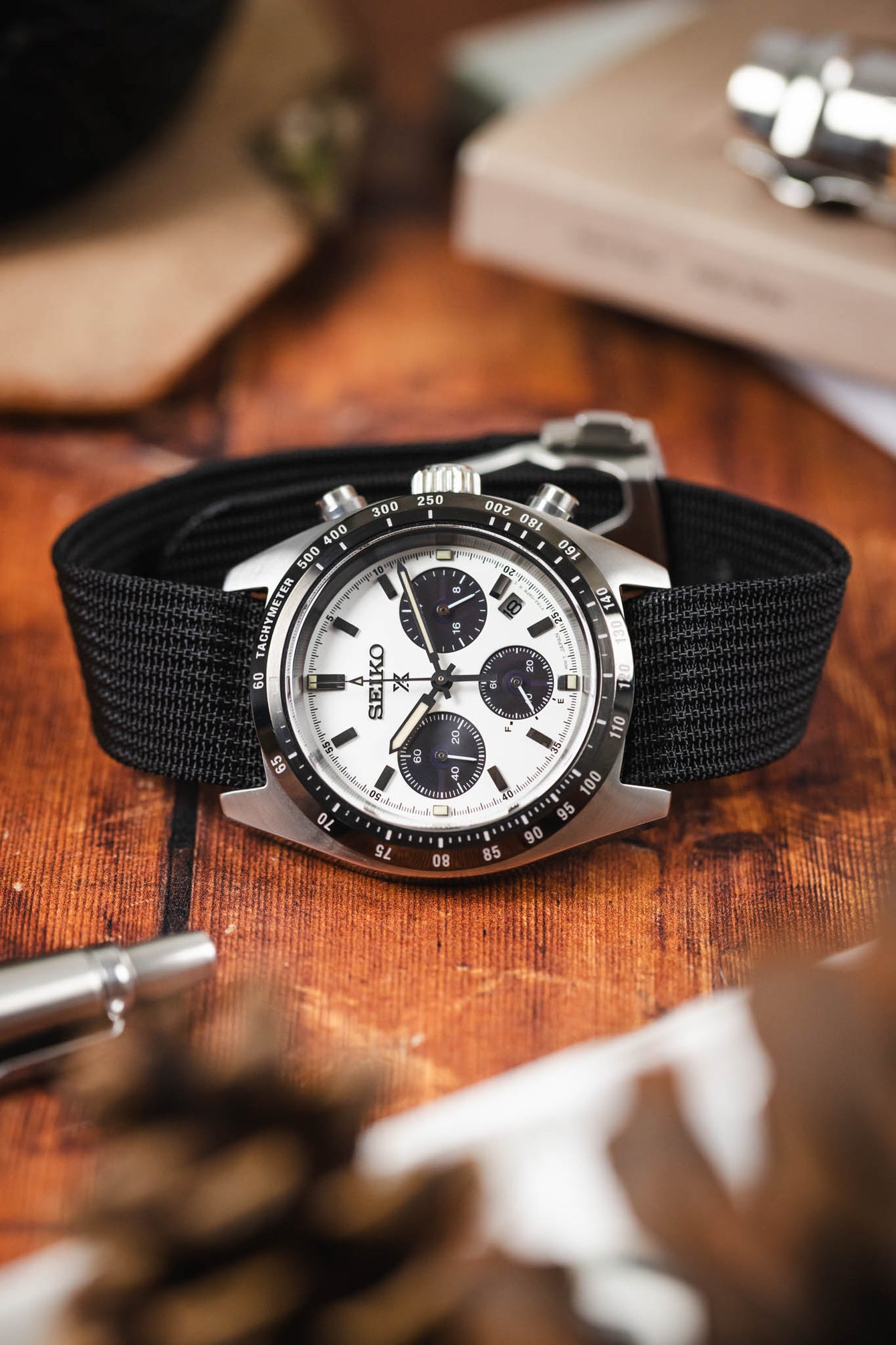 Close-up of a stainless steel Omega Speedmaster chronograph with a white dial and black tachymeter bezel, fitted on a burgundy fabric strap, resting on a wooden surface with a book, a metal tool, and softly blurred greenery in the foreground.