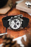 Close-up of a stainless steel Omega Speedmaster chronograph with a white dial and black tachymeter bezel, fitted on a burgundy fabric strap, resting on a wooden surface with a book, a metal tool, and softly blurred greenery in the foreground.