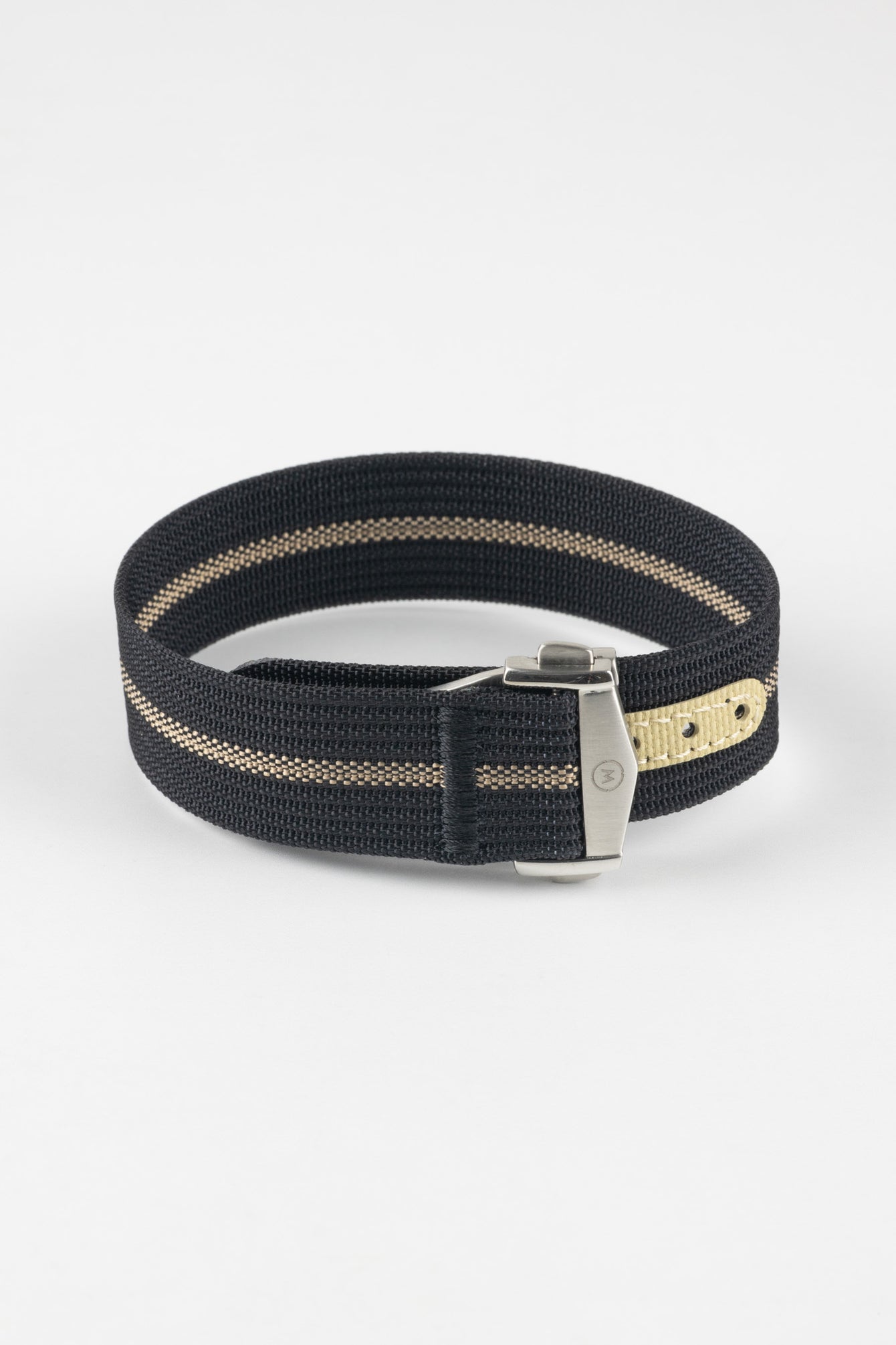 Close-up of a black ribbed nylon watch strap with a central cream stripe, arranged in a loop on a light grey background. The strap features a brushed silver deployant clasp with an engraved logo and a cream-colored reinforced section with evenly spaced adjustment holes positioned near the clasp.