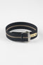 Close-up of a black ribbed nylon watch strap with a central cream stripe, arranged in a loop on a light grey background. The strap features a brushed silver deployant clasp with an engraved logo and a cream-colored reinforced section with evenly spaced adjustment holes positioned near the clasp.
