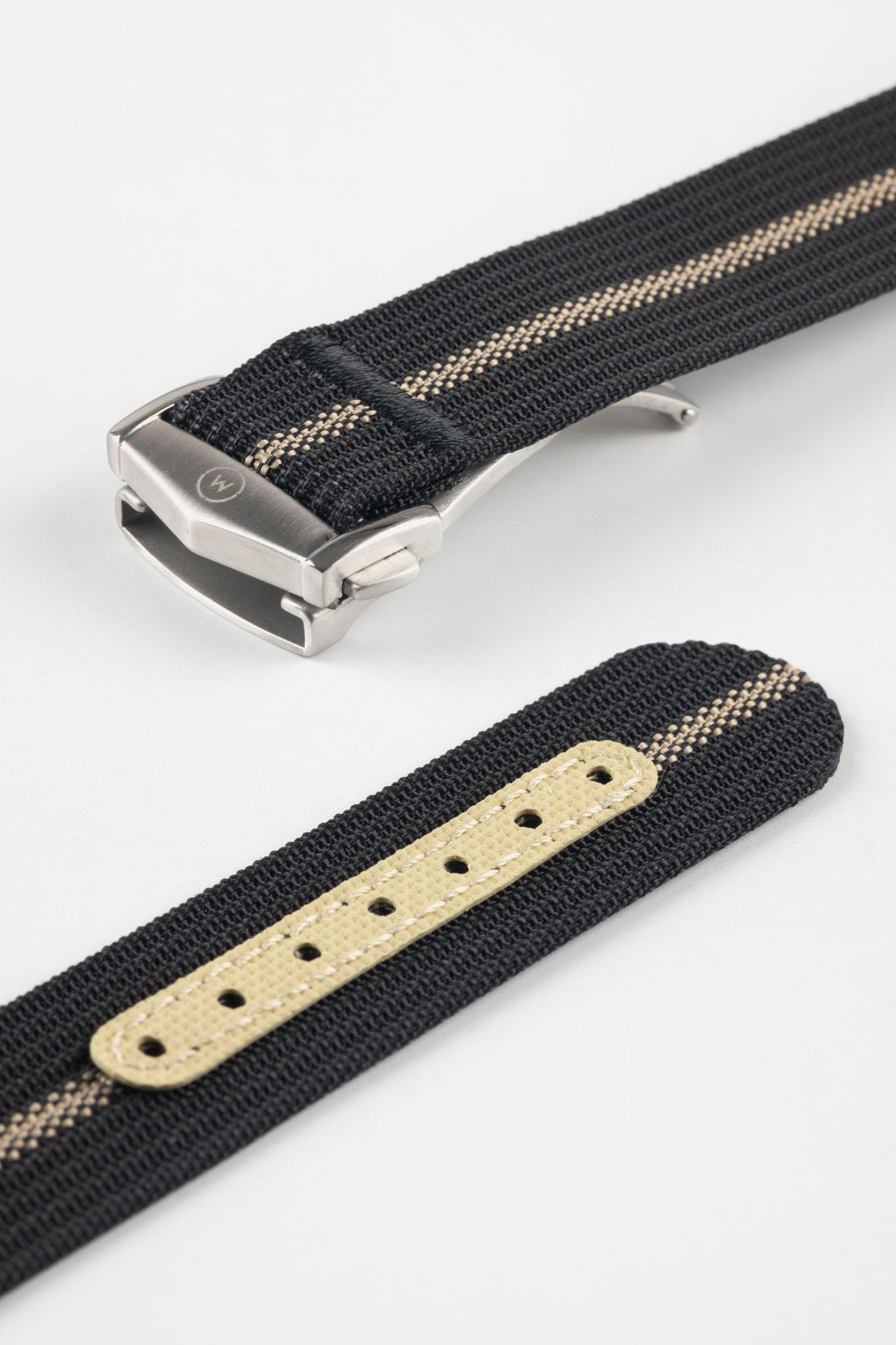 Angled close-up of a black ribbed nylon watch strap with a central cream stripe, featuring a brushed silver deployant clasp with an engraved logo. A separate strap section shows a cream-colored reinforced panel with evenly spaced adjustment holes, all set against a clean light grey background.