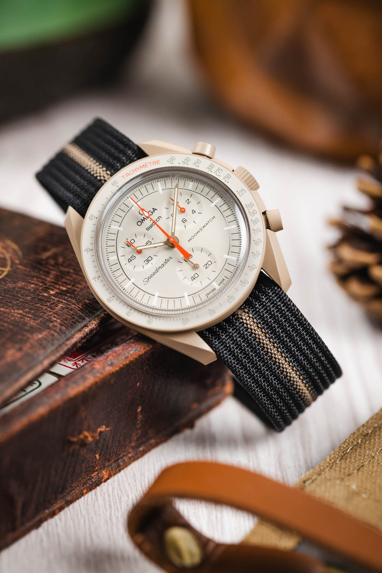 Close-up of a light beige chronograph-style wristwatch with a tachymeter bezel and white dial, featuring subdials and a bright orange seconds hand, resting against a worn wooden surface. The watch is fitted with a black ribbed nylon strap with a central cream stripe. Surrounding elements include blurred leather accessories and warm-toned objects, creating a rustic lifestyle setting.