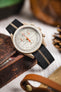 Close-up of a light beige chronograph-style wristwatch with a tachymeter bezel and white dial, featuring subdials and a bright orange seconds hand, resting against a worn wooden surface. The watch is fitted with a black ribbed nylon strap with a central cream stripe. Surrounding elements include blurred leather accessories and warm-toned objects, creating a rustic lifestyle setting.