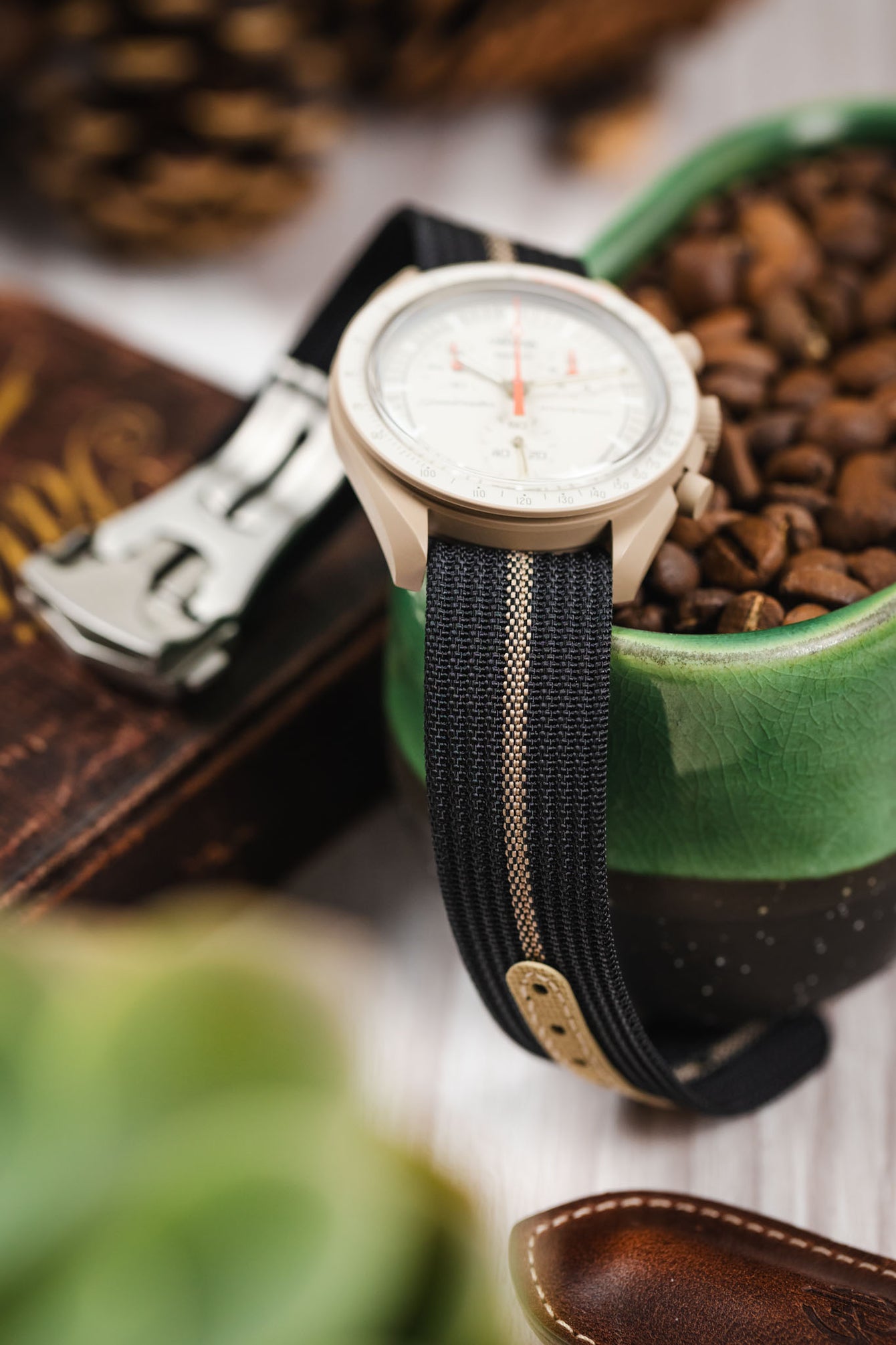 Close-up of a light beige chronograph-style watch positioned on the edge of a green ceramic dish filled with coffee beans. The watch features a white dial with subdials and an orange seconds hand, and is paired with a black ribbed nylon strap with a central cream stripe. In the softly blurred background are warm-toned elements including a wooden object and pinecones, creating a rustic, lifestyle setting.