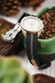 Close-up of a light beige chronograph-style watch positioned on the edge of a green ceramic dish filled with coffee beans. The watch features a white dial with subdials and an orange seconds hand, and is paired with a black ribbed nylon strap with a central cream stripe. In the softly blurred background are warm-toned elements including a wooden object and pinecones, creating a rustic, lifestyle setting.