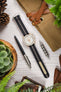 Styled flat lay of a light beige chronograph-style watch with a white dial and orange seconds hand, fitted on a black ribbed nylon strap with a central cream stripe. The watch is arranged on a light wooden surface alongside lifestyle accessories including a tan canvas tool roll with leather straps, a metal watch tool, a pen, a pinecone, and green houseplants. The scene features warm tones and soft natural textures, creating a rustic, outdoors-inspired aesthetic.