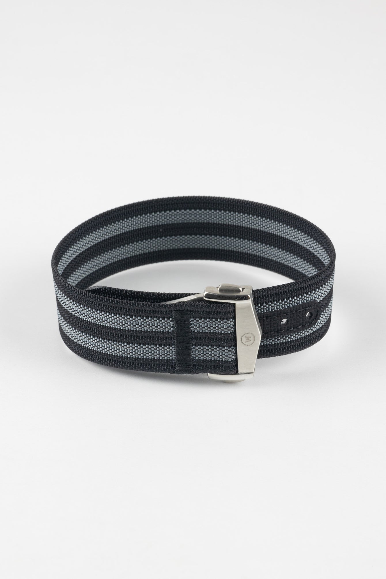 Close-up of a black ribbed nylon watch strap with grey and white striped detailing, arranged in a loop on a light grey background. The strap features a brushed silver deployant clasp with an engraved logo and a dark reinforced section with adjustment holes positioned near the clasp.