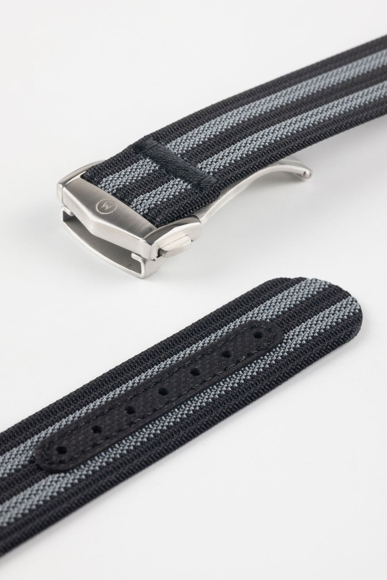 Angled close-up of a black ribbed nylon watch strap with alternating grey stripes, featuring a brushed silver deployant clasp with an engraved logo. A separate strap section shows a dark reinforced panel with evenly spaced adjustment holes, all displayed on a light grey background.