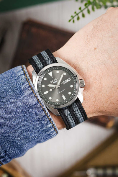 Close-up of a wrist wearing a Seiko 5 automatic watch with a grey sunburst dial, round and triangular hour markers, and a day-date display at the 3 o’clock position. The watch is paired with a black ribbed nylon strap featuring grey and white striped detailing. A stainless steel case and crown are visible, alongside a blue denim sleeve. The background is softly blurred with warm tones and hints of greenery, creating a casual lifestyle setting.