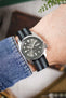 Close-up of a wrist wearing a Seiko 5 automatic watch with a grey sunburst dial, round and triangular hour markers, and a day-date display at the 3 o’clock position. The watch is paired with a black ribbed nylon strap featuring grey and white striped detailing. A stainless steel case and crown are visible, alongside a blue denim sleeve. The background is softly blurred with warm tones and hints of greenery, creating a casual lifestyle setting.