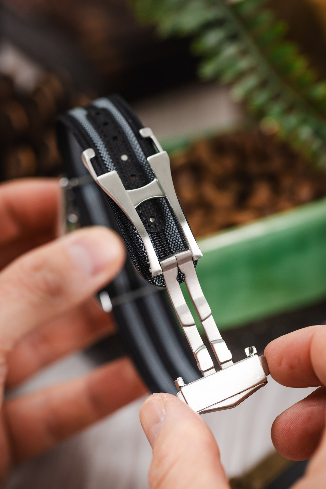 Close-up of a black ribbed nylon watch strap with grey striped detailing being held by fingers, highlighting a brushed silver deployant clasp in an open position. The clasp mechanism and hinge are clearly visible, along with a dark reinforced section featuring adjustment holes. The background is softly blurred with warm tones, including coffee beans and greenery, creating a lifestyle setting.