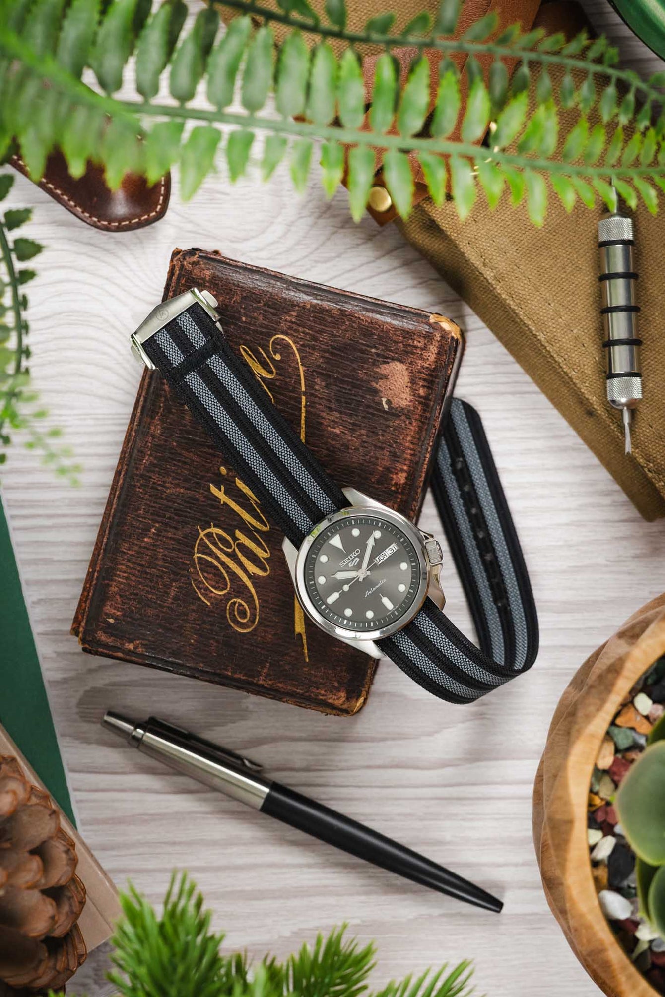 Styled flat lay of a Seiko automatic watch with a grey sunburst dial and day-date display, paired with a black ribbed nylon strap featuring grey striped detailing. The watch rests on a worn brown book while surrounded by lifestyle items including a metal watch tool, a pen, a tan canvas roll, and green foliage. The scene is arranged on a light wooden surface with warm tones and a natural, outdoors-inspired aesthetic.