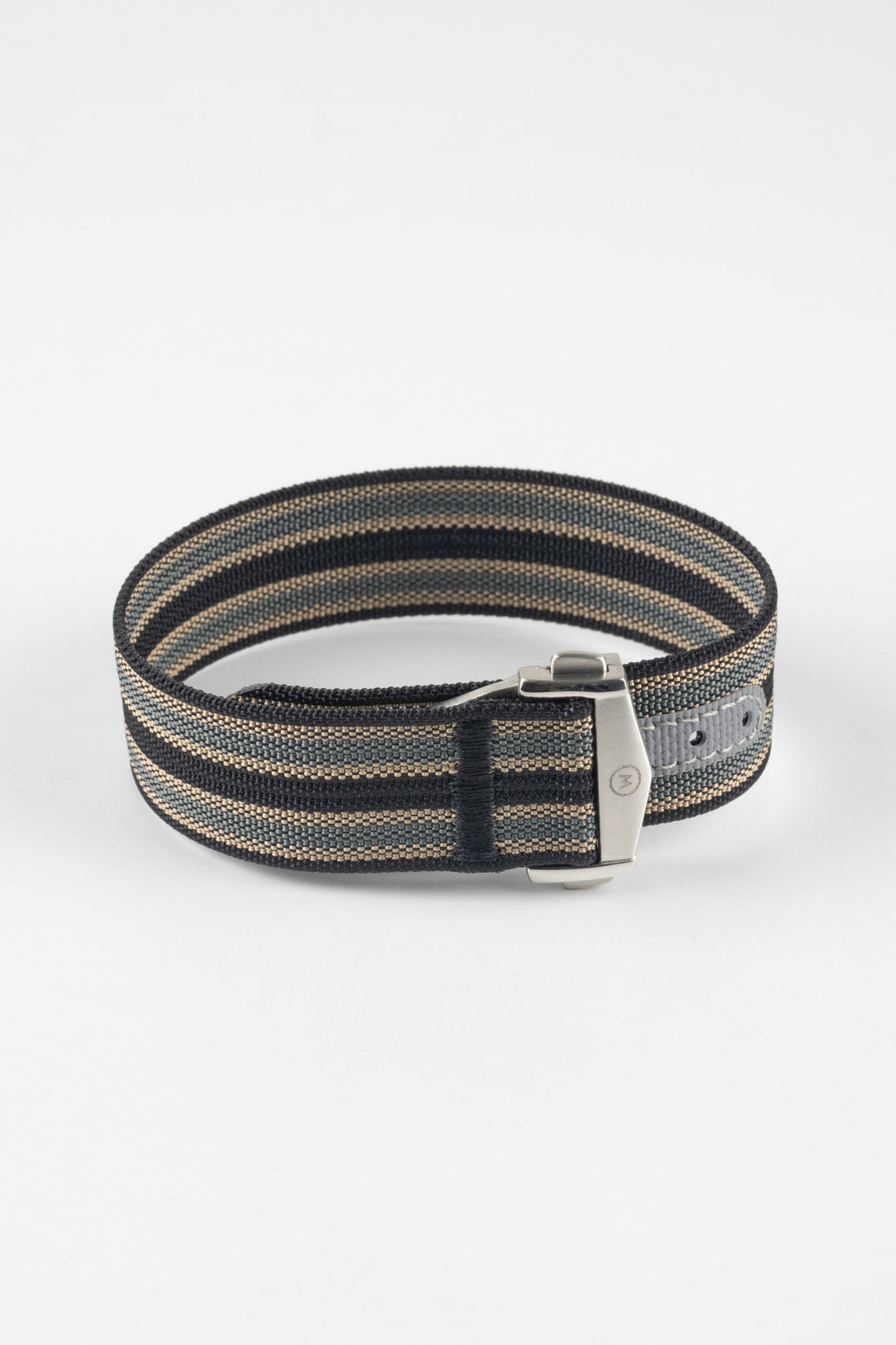 Close-up of a striped ribbed nylon watch strap in black, grey, and cream tones, shown horizontally on a light grey background, featuring a brushed silver deployant clasp with an engraved logo and a grey reinforced section with adjustment holes.