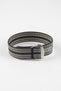 Close-up of a striped ribbed nylon watch strap in black, grey, and cream tones, shown horizontally on a light grey background, featuring a brushed silver deployant clasp with an engraved logo and a grey reinforced section with adjustment holes.