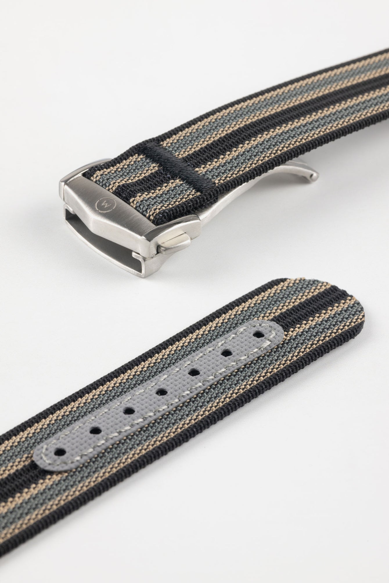 Angled close-up of a striped ribbed nylon watch strap in black, grey, and cream tones, featuring a brushed silver deployant clasp with an engraved logo, alongside a separate strap section with a grey reinforced panel and adjustment holes, all set against a light grey background.