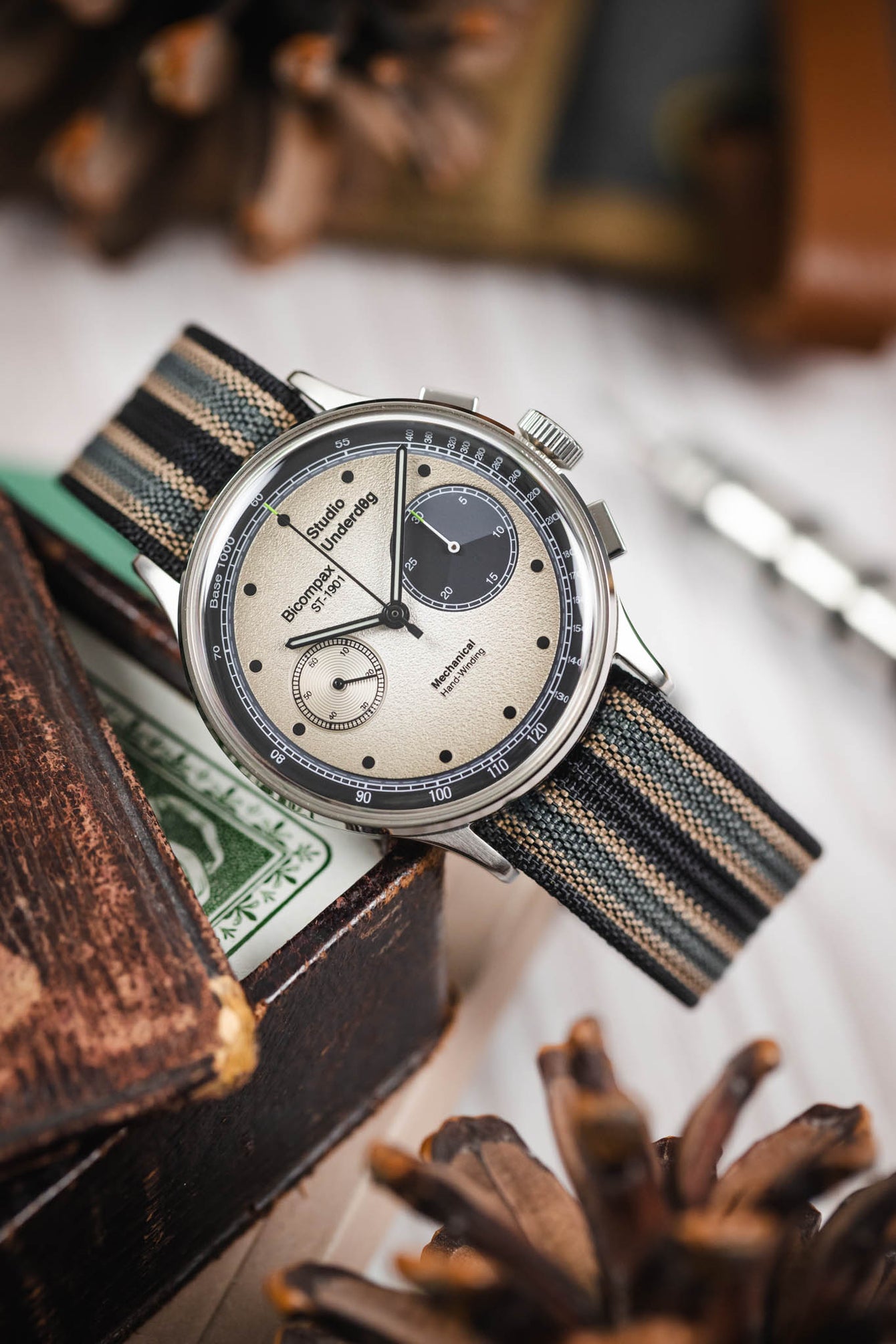 Close-up of a chronograph-style watch with a cream dial and black subdials, fitted with a striped ribbed nylon strap in black, grey, and cream tones, resting across a worn wooden surface and small decorative objects. The polished silver case, crown, and pushers are visible, and the softly blurred background includes neutral indoor elements.