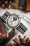 Close-up of a chronograph-style watch with a cream dial and black subdials, fitted with a striped ribbed nylon strap in black, grey, and cream tones, resting across a worn wooden surface and small decorative objects. The polished silver case, crown, and pushers are visible, and the softly blurred background includes neutral indoor elements.