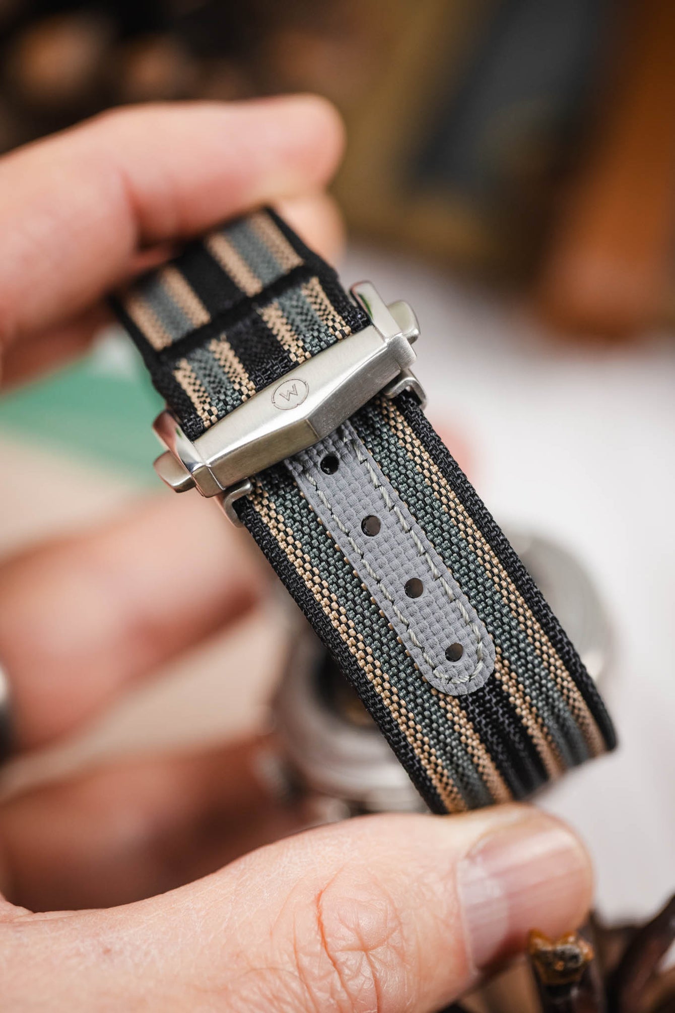 Close-up of a striped ribbed nylon watch strap in black, grey, and cream tones being held in a hand, highlighting a brushed silver deployant clasp with an engraved logo. A grey reinforced section with adjustment holes is visible along the strap, with a softly blurred indoor background featuring warm tones and decorative objects.