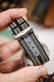 Close-up of a striped ribbed nylon watch strap in black, grey, and cream tones being held in a hand, highlighting a brushed silver deployant clasp with an engraved logo. A grey reinforced section with adjustment holes is visible along the strap, with a softly blurred indoor background featuring warm tones and decorative objects.