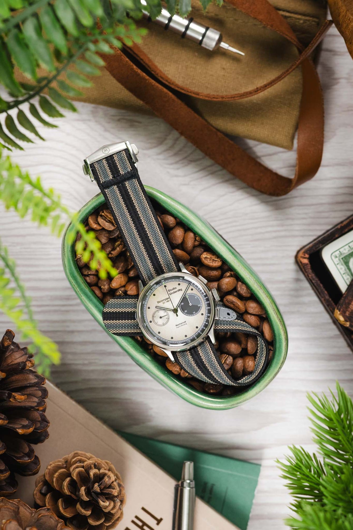 Styled flat lay of a chronograph-style wristwatch with a cream dial and black subdials, fitted with a striped ribbed nylon strap in black, grey, and cream tones, resting inside a green dish filled with coffee beans. Surrounding elements include pinecones, greenery, a tan cloth pouch with a watch tool, and a white wooden surface, creating a warm, rustic lifestyle setting.