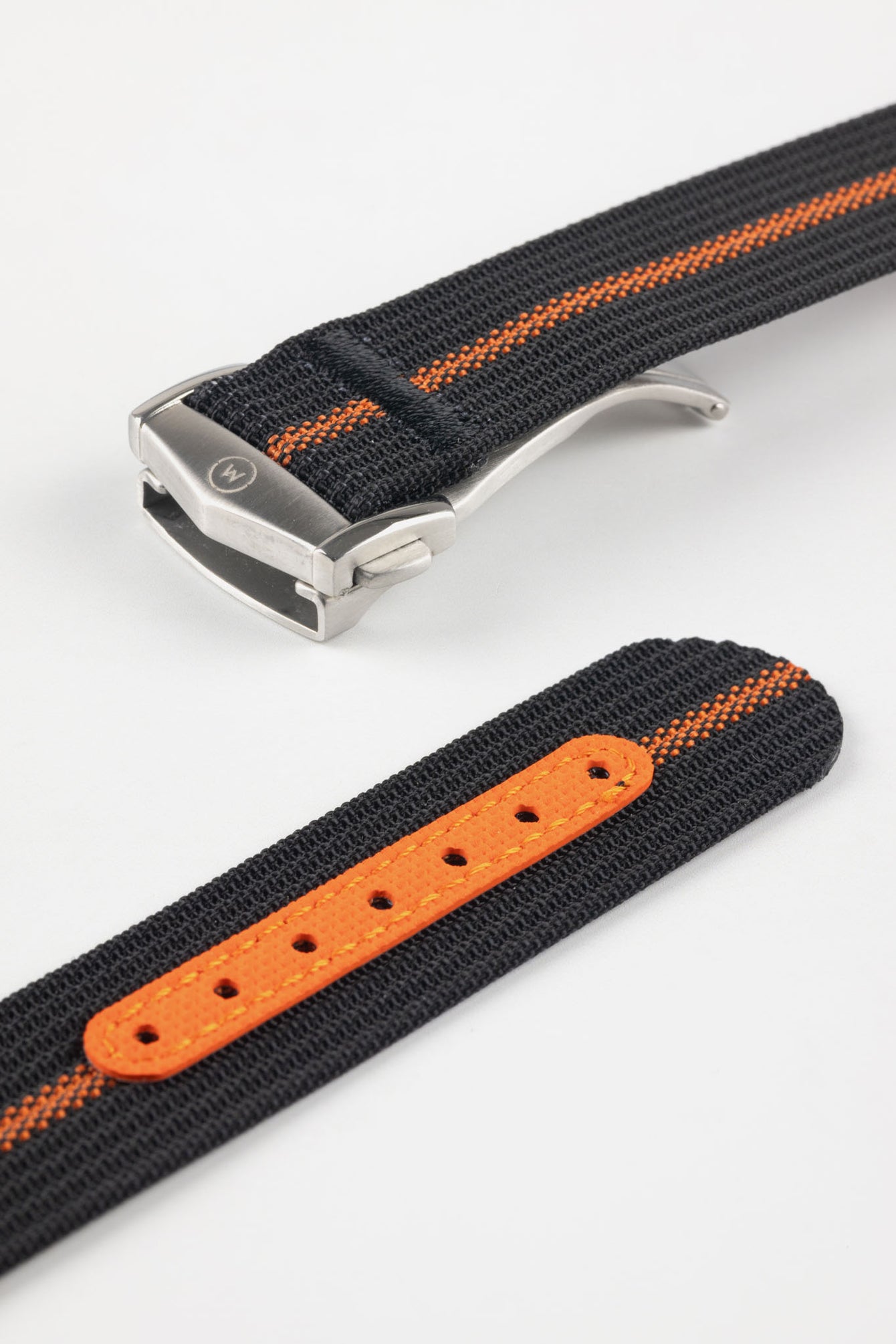 Angled close-up of a black ribbed nylon watch strap with a central orange stripe, featuring a brushed silver deployant clasp with an engraved logo. A separate section of the strap shows a bright orange reinforced panel with evenly spaced adjustment holes, all displayed on a light grey background.