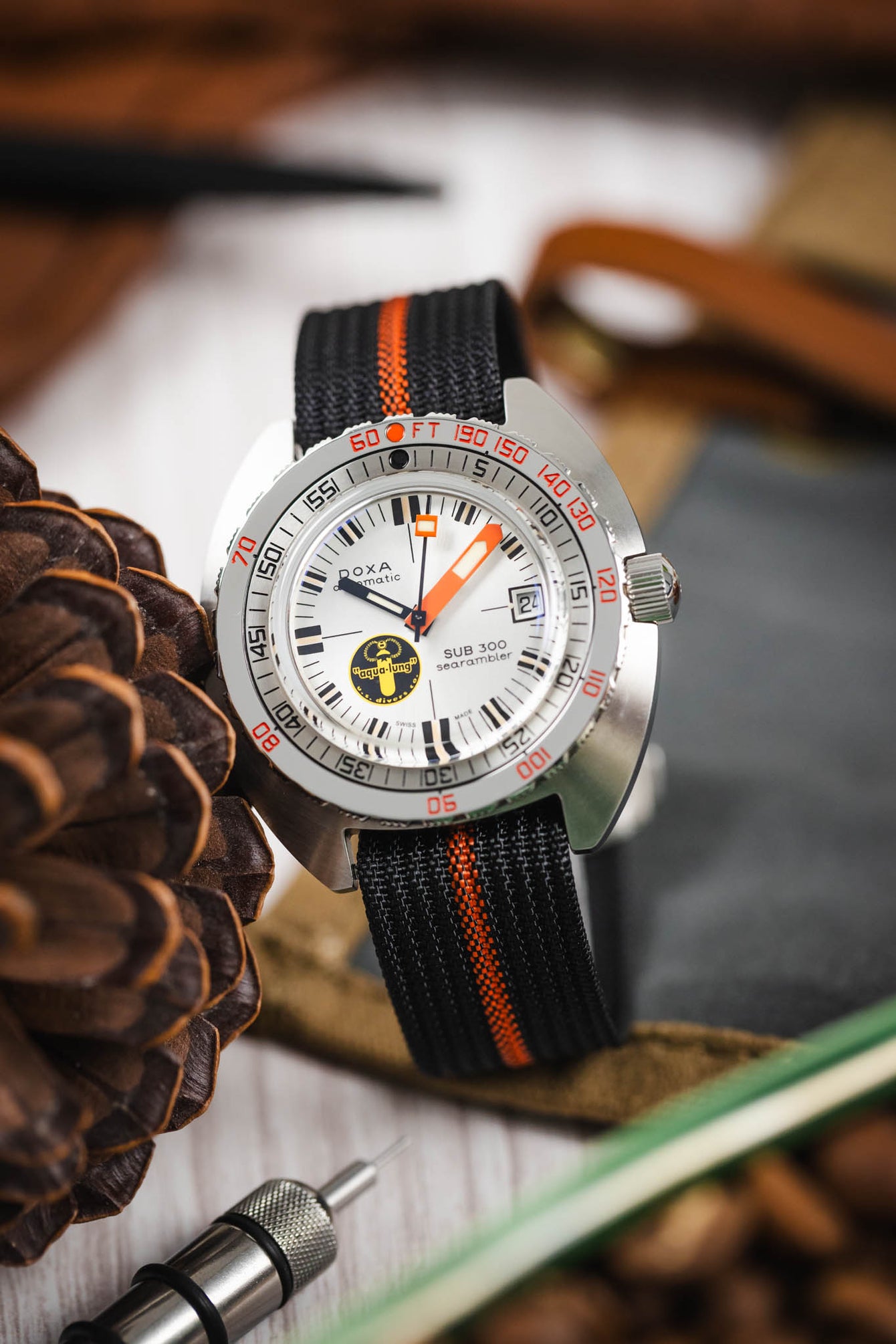 Close-up of a dive-style watch with a white dial, black hour markers, and orange accents, including a bold orange minute hand and bezel markings. The watch is fitted with a black ribbed nylon strap featuring a central orange stripe and is displayed among lifestyle props such as a pinecone, leather items, and a metal watch tool on a wooden surface, with a softly blurred warm-toned background.