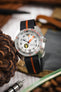 Close-up of a dive-style watch with a white dial, black hour markers, and orange accents, including a bold orange minute hand and bezel markings. The watch is fitted with a black ribbed nylon strap featuring a central orange stripe and is displayed among lifestyle props such as a pinecone, leather items, and a metal watch tool on a wooden surface, with a softly blurred warm-toned background.