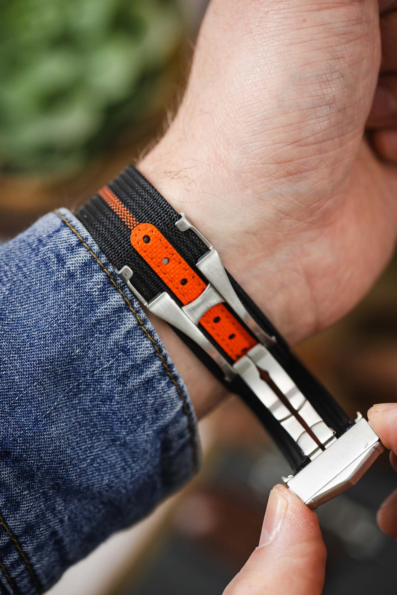 Close-up of a wrist wearing a black ribbed nylon watch strap with a central orange stripe, showing the underside with a bright orange reinforced section and adjustment holes. A brushed silver deployant clasp is being fastened, highlighting the mechanism and fit against a blue denim sleeve, with a softly blurred warm-toned background.
