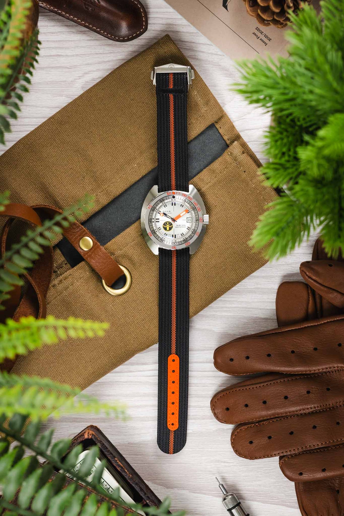 Styled flat lay of a dive-style watch with a white dial and orange accents on a black ribbed nylon strap with a central orange stripe, arranged vertically on a tan canvas pouch. Surrounding objects include brown leather gloves, a leather strap with brass hardware, a pen, a metal watch tool, pinecones, and green foliage, creating a warm, outdoors-inspired setting on a light wooden surface.