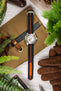 Styled flat lay of a dive-style watch with a white dial and orange accents on a black ribbed nylon strap with a central orange stripe, arranged vertically on a tan canvas pouch. Surrounding objects include brown leather gloves, a leather strap with brass hardware, a pen, a metal watch tool, pinecones, and green foliage, creating a warm, outdoors-inspired setting on a light wooden surface.