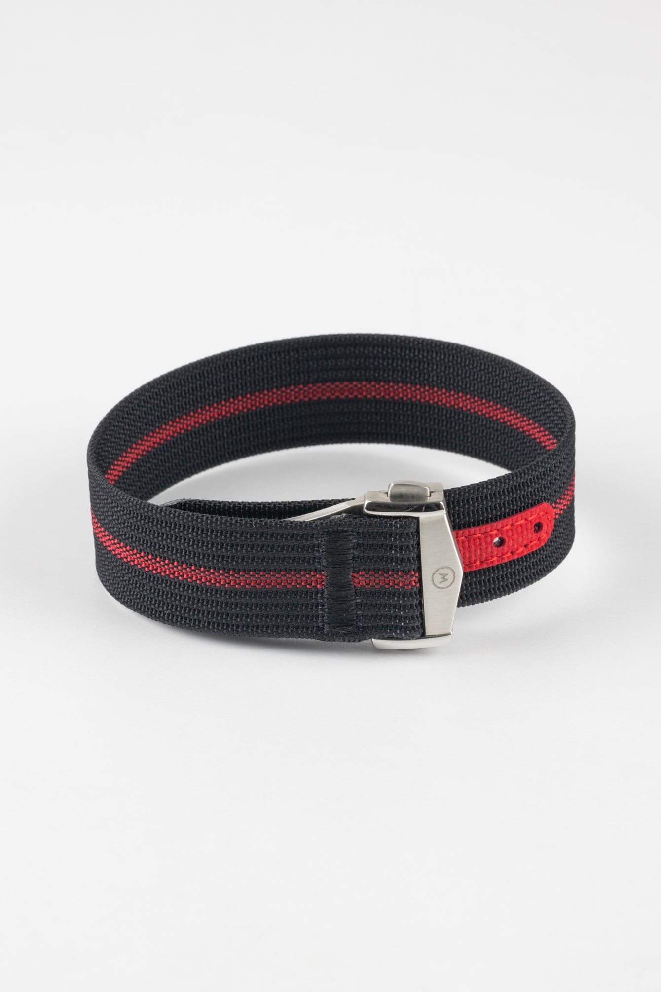 Close-up of a black ribbed nylon watch strap with a central red stripe, arranged in a loop on a light grey background. The strap features a brushed silver deployant clasp with an engraved logo and a red reinforced section with evenly spaced adjustment holes near the clasp.