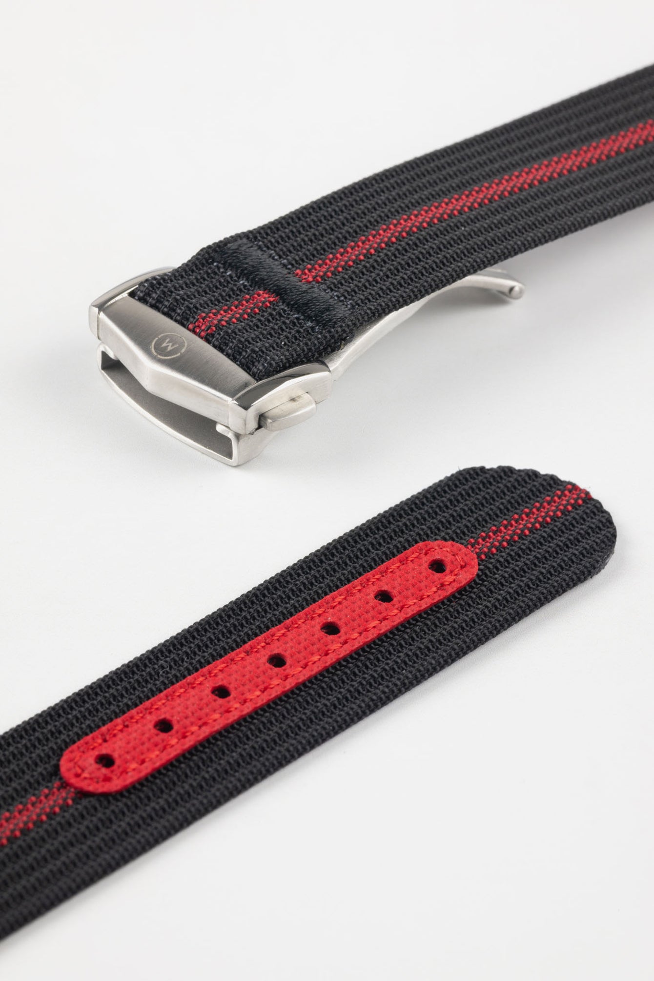 Angled close-up of a black ribbed nylon watch strap with a central red stripe, featuring a brushed silver deployant clasp with an engraved logo. A separate strap section displays a red reinforced panel with evenly spaced adjustment holes, all set against a light grey background.