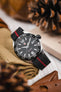 Close-up of a TAG Heuer Formula 1 watch with a black dial and bold white numeral markers, resting on a folded tan canvas pouch. The watch is fitted with a black ribbed nylon strap featuring a central red stripe. Surrounding elements include a pinecone, leather accessories, and blurred warm-toned objects, creating a rustic, lifestyle product setting.