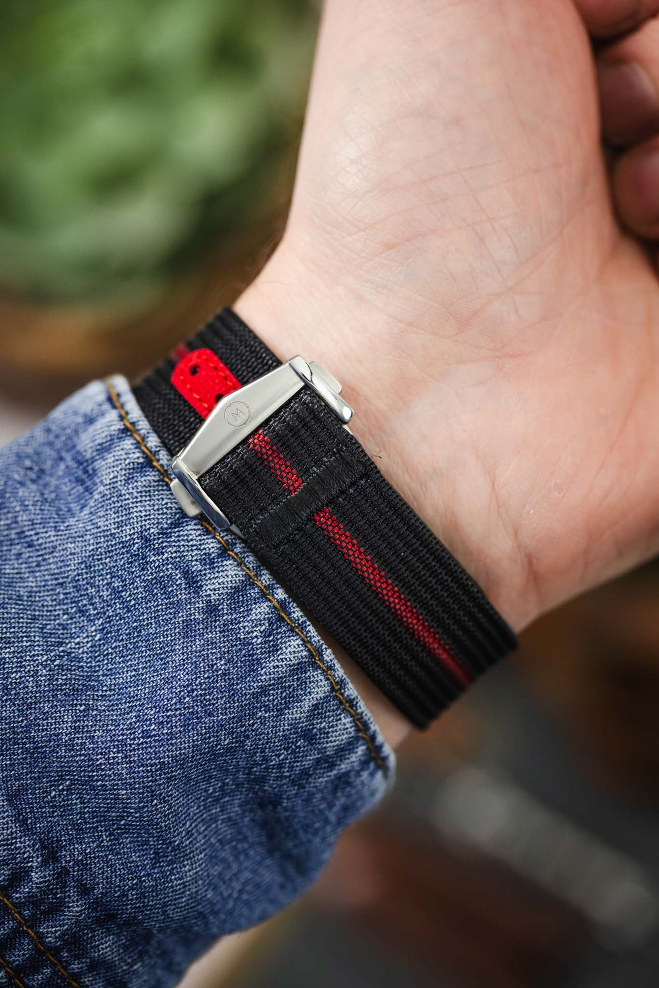 Close-up of the underside of a wrist wearing a black ribbed nylon watch strap with a central red stripe. The strap features a red reinforced section with adjustment holes and a brushed silver deployant clasp with an engraved logo. The strap is shown against a blue denim sleeve, with a softly blurred background of green foliage and warm tones.