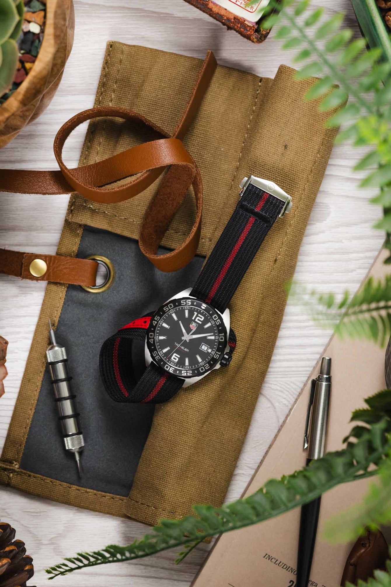 Styled flat lay of a TAG Heuer Formula 1 watch with a black dial and white markers, fitted with a black ribbed nylon strap featuring a central red stripe. The watch is arranged on a tan canvas tool roll alongside a brown leather strap with brass hardware, a metal watch tool, a pen, pinecones, and green foliage, all set on a light wooden surface for a warm, outdoor-inspired lifestyle scene.