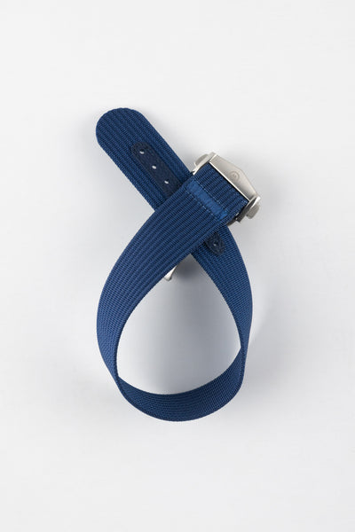 A top-down shot of a blue ribbed-nylon watch strap coiled into a loop against a white background, featuring a brushed stainless steel buckle with a circular logo.
