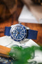 Close-up of a stainless steel Tudor dive watch with a blue dial and matching blue fabric strap, resting on a wooden surface and partially supported by a ceramic object with an octopus illustration, with a softly blurred background and green plant leaves in the foreground.