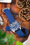 Close-up of a stainless steel Tudor dive watch with a blue dial and matching blue fabric strap, resting on a wooden surface beside pinecones, with soft green plant leaves in the foreground and a blurred background of everyday items.