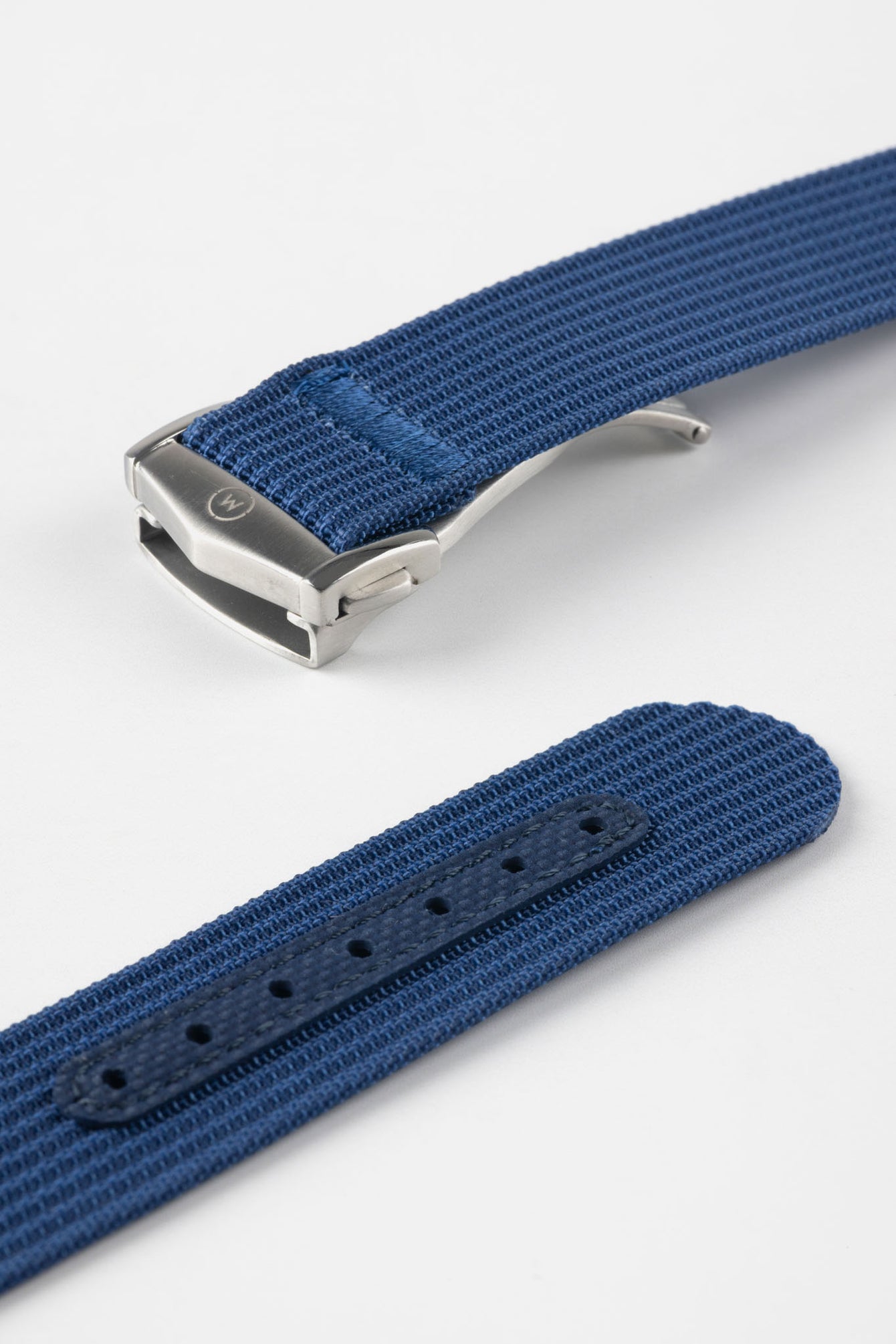 A close-up, angled shot of a blue ribbed-nylon watch strap showing both the brushed stainless steel deployment buckle and the adjustment holes. The image highlights the fine woven texture of the fabric and the precision stitching near the metal clasp.