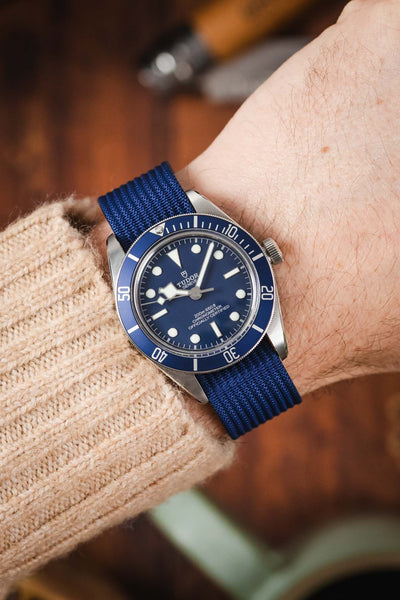 A close-up shot of a person's wrist wearing a Tudor BB58 watch with a wlue dial. The watch is paired with a blue ribbed-nylon strap and is worn over the sleeve of a beige cable-knit sweater, creating a cozy, outdoor-inspired look.