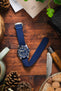 A top-down lifestyle shot of a Tudor Black Bay 58 chronograph watch with a white dial and black sub-dials, fitted on a blue ribbed-nylon strap. The watch rests on a rustic leather pouch atop a wooden surface, surrounded by pine cones, an Opinel folding knife, and green foliage.