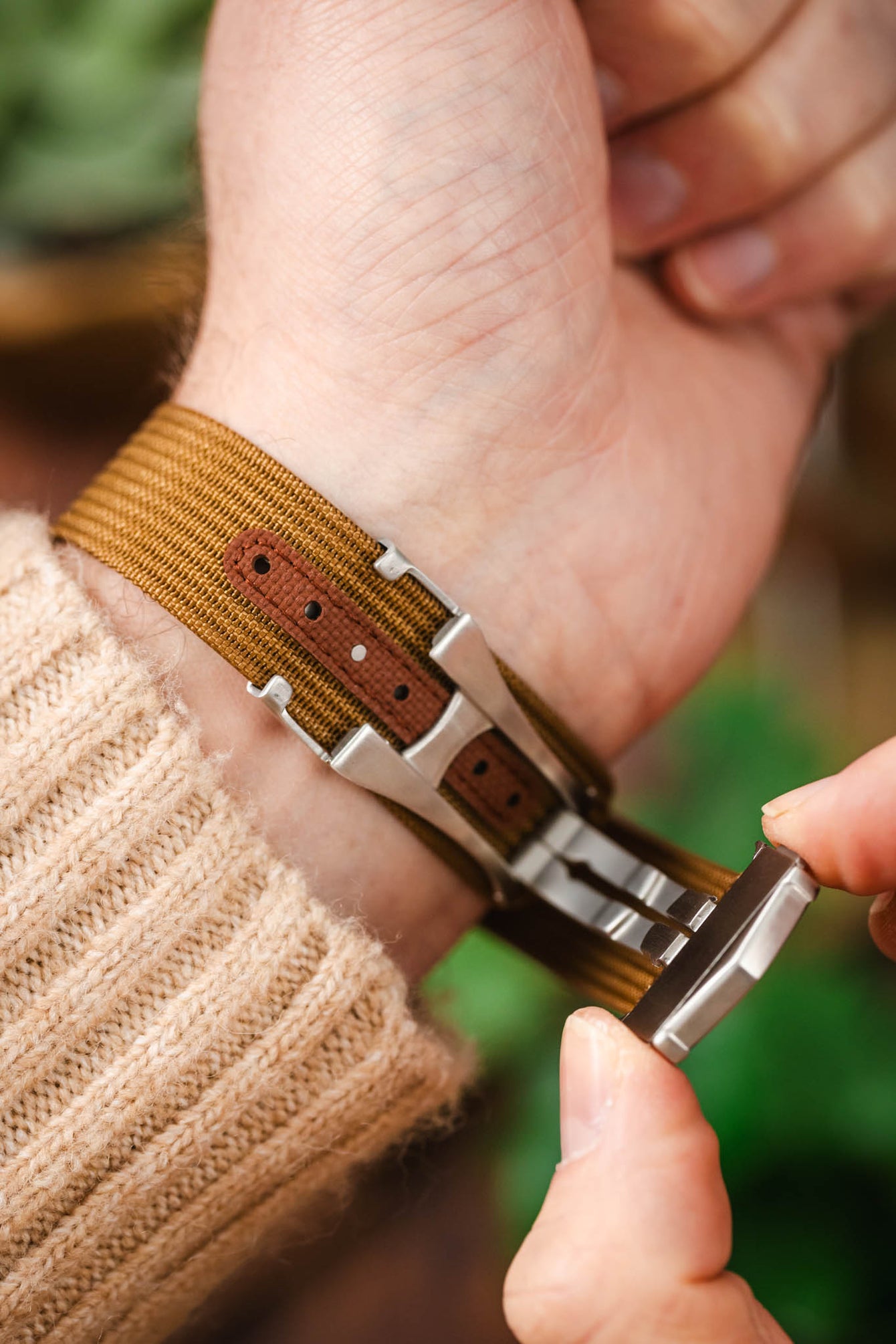 Close-up of a wrist wearing a brown ribbed nylon watch strap with a darker reinforced section and adjustment holes, as a brushed silver deployant clasp is being fastened against a blurred outdoor background.