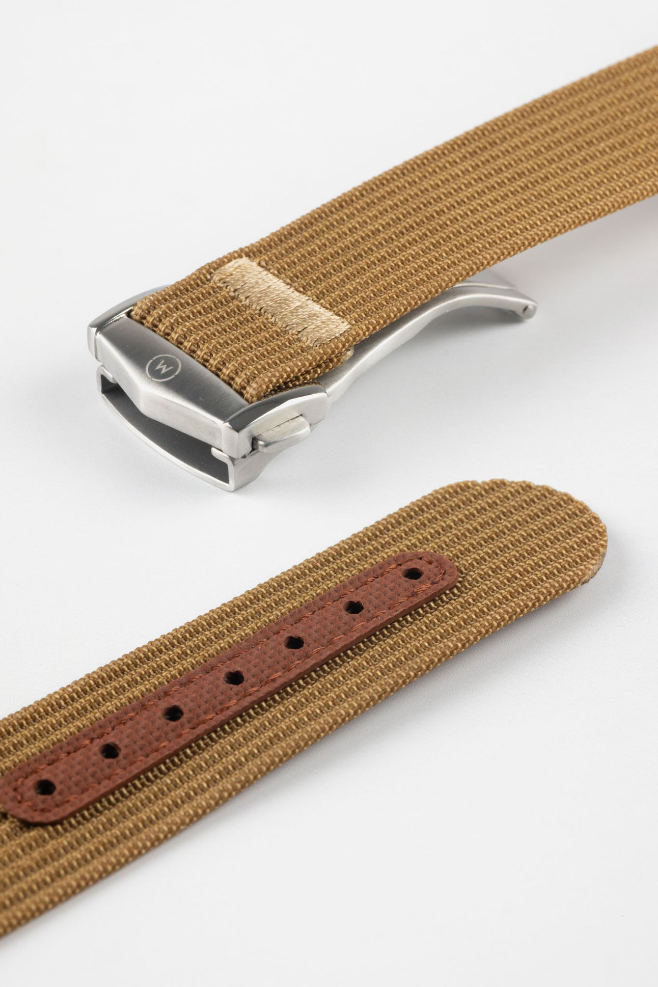 A close-up, angled shot of a brown ribbed-nylon watch strap showing both the brushed stainless steel deployment buckle and the adjustment holes. The image highlights the fine woven texture of the fabric and the precision stitching near the metal clasp.