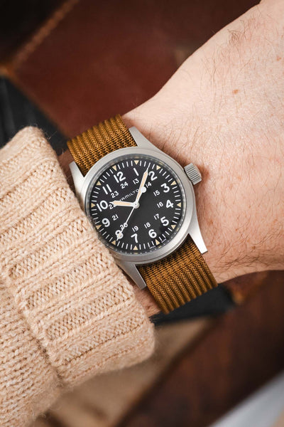 A close-up shot of a person's wrist wearing a Hamilton Khaki Field timer watch with a pure black dial. The watch is paired with a brown ribbed-nylon strap and is worn over the sleeve of a beige cable-knit sweater, creating a cozy, outdoor-inspired look.