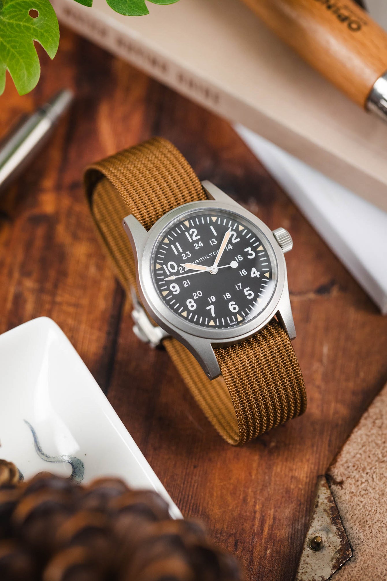 Field-style wristwatch with a black dial and white numerals on a brown ribbed nylon strap, displayed on a wooden surface alongside a pen, book, and greenery in a warm lifestyle setting.