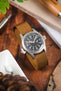 Field-style wristwatch with a black dial and white numerals on a brown ribbed nylon strap, displayed on a wooden surface alongside a pen, book, and greenery in a warm lifestyle setting.