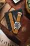 A top-down lifestyle shot of a Hamilton  Khaki Field chronograph watch with a white dial and black sub-dials, fitted on a brown ribbed-nylon strap. The watch rests on a rustic leather pouch atop a wooden surface, surrounded by pine cones, an Opinel folding knife, and green foliage.