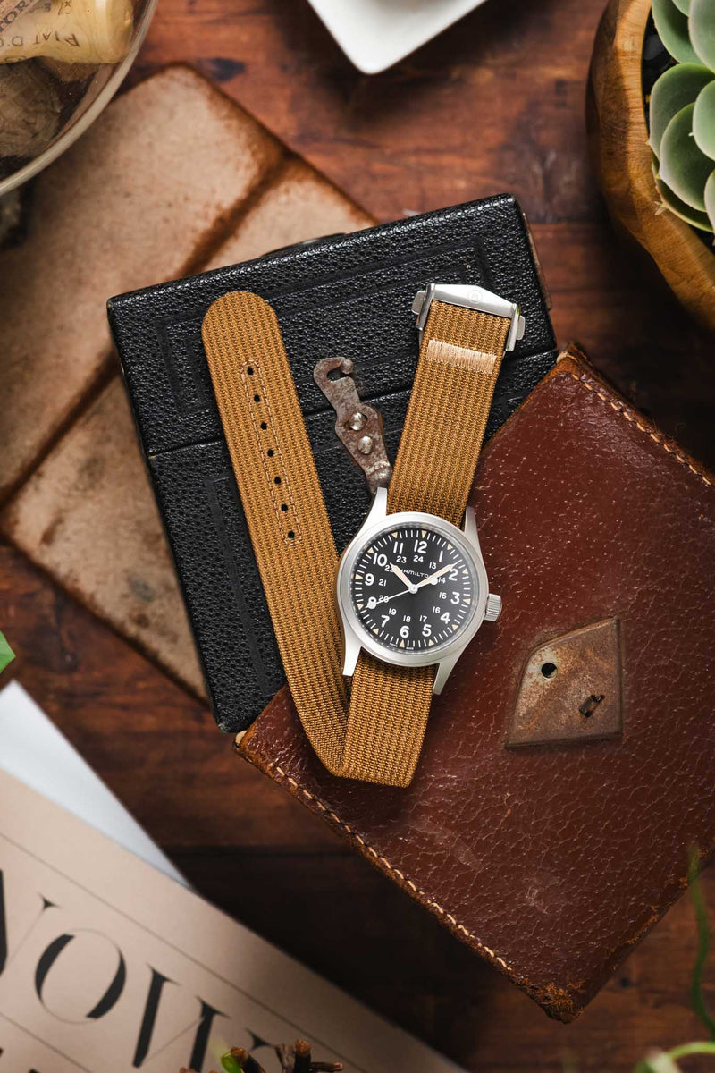 A top-down lifestyle shot of a Hamilton  Khaki Field chronograph watch with a white dial and black sub-dials, fitted on a brown ribbed-nylon strap. The watch rests on a rustic leather pouch atop a wooden surface, surrounded by pine cones, an Opinel folding knife, and green foliage.