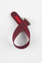 A top-down shot of a burgundy ribbed-nylon watch strap coiled into a loop against a white background, featuring a brushed stainless steel buckle with a circular logo.