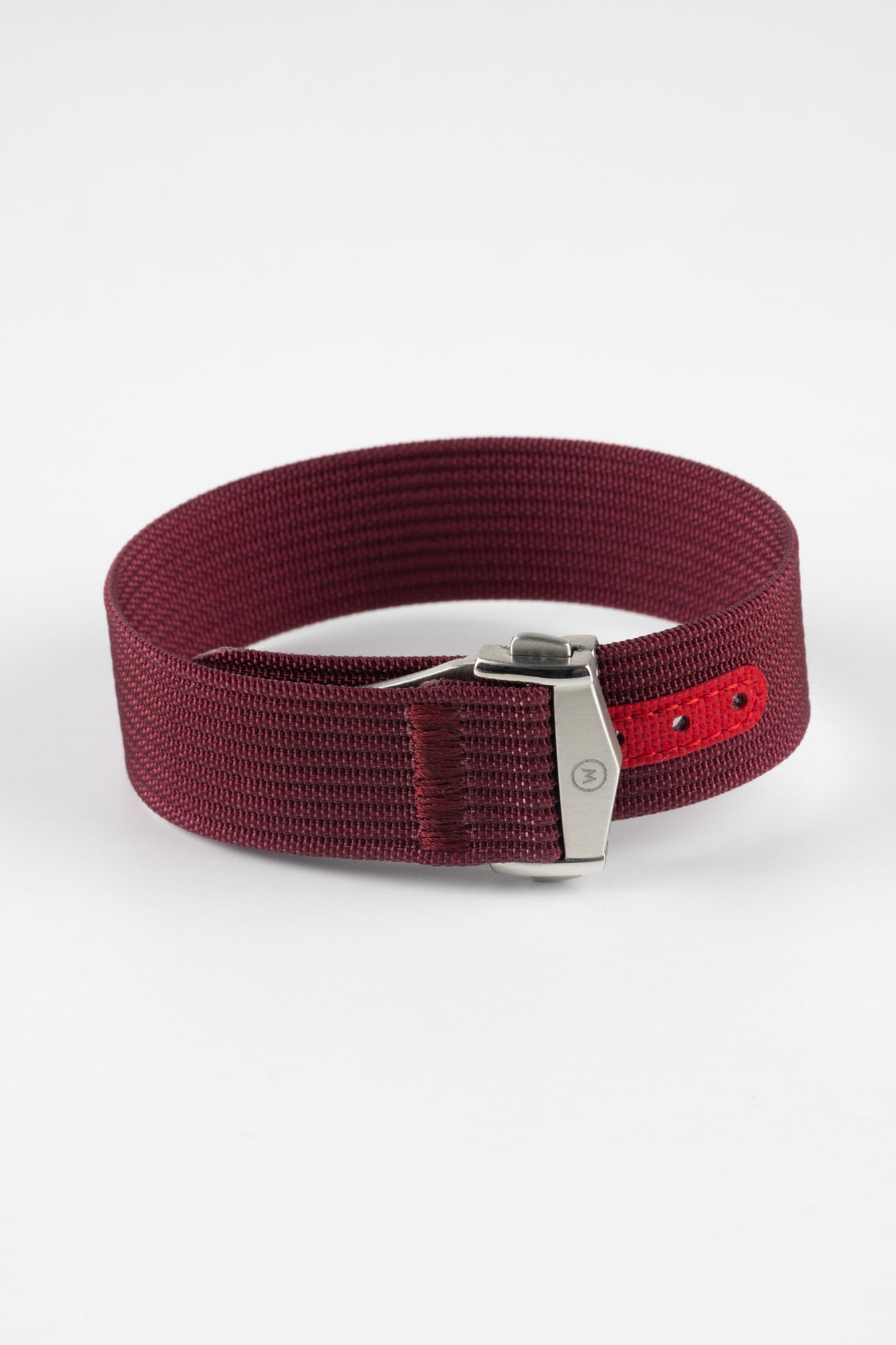 A three-quarter view of a burgundy ribbed-nylon watch strap fastened into a loop, highlighting the brushed metal buckle and woven texture on a white background.